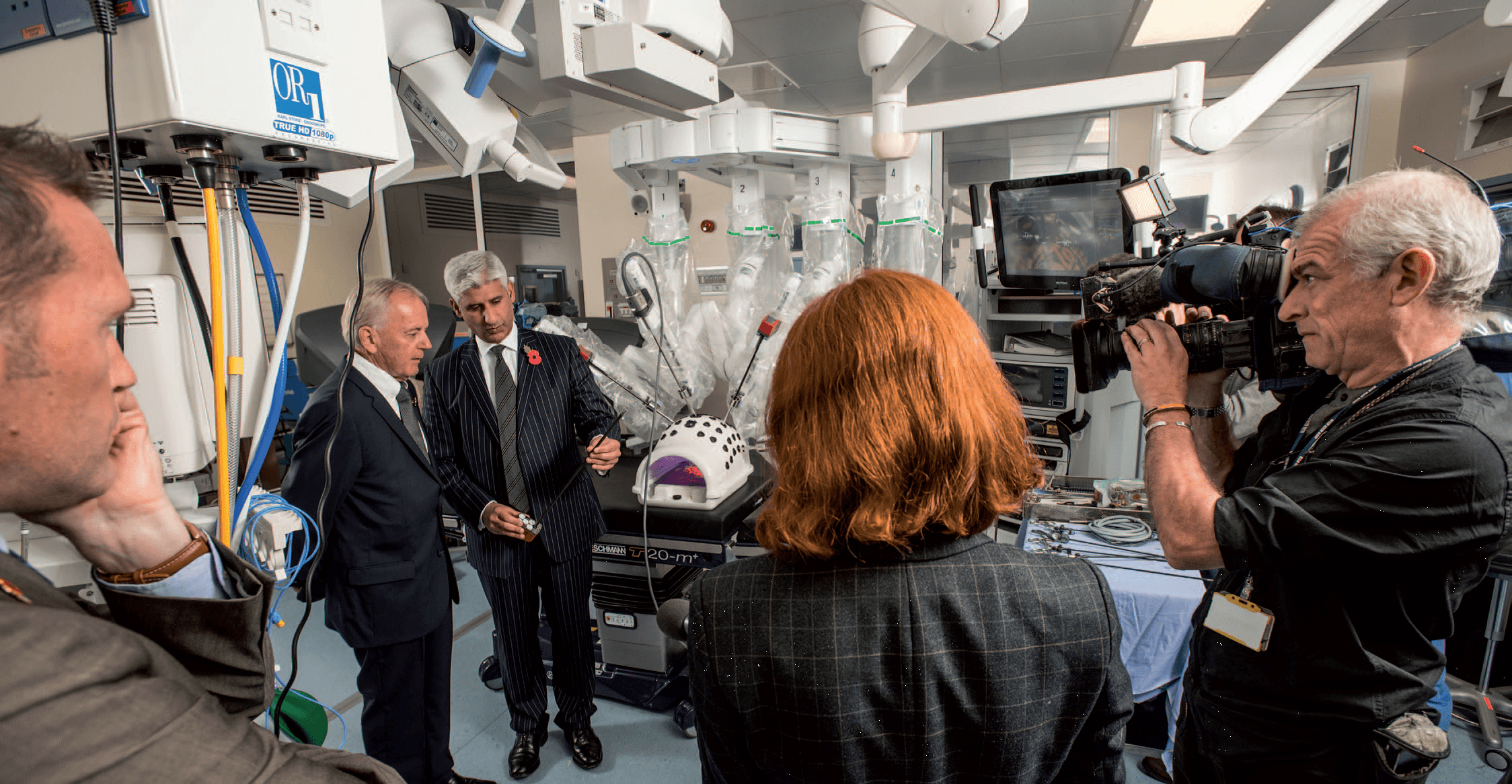 Professor Tas Qureshi and Robert Braithwaite being filmed by a press crew beside the da Vinci robotic system in theatre.